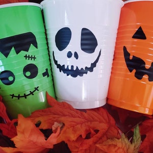 May include: Three Halloween-themed plastic cups: green Frankenstein, white ghost, and orange jack-o'-lantern. Each cup features a black graphic face design. The cups are set on a bed of orange and red autumn leaves.