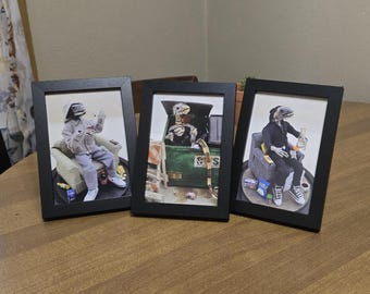 May include: Three framed art prints featuring anthropomorphic lizard figures in various settings. One figure is seated, another is in a dumpster, and the third is seated, all with detailed surroundings and black frames. The art has a unique, quirky style.