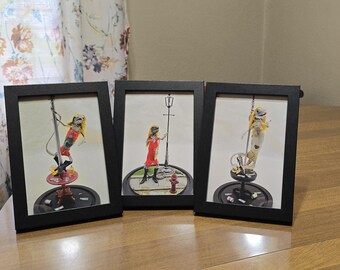 May include: Three framed art prints. Each features a skeleton figure in a red dress and black boots. One is on a pole, another by a street lamp, and the third on a pole. The frames are black, and the background is white.
