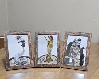 May include: Three framed pictures of lizards dressed as cowboys. The lizards are wearing hats, vests, and holding guns. The frames are made of wood and have a rustic look.