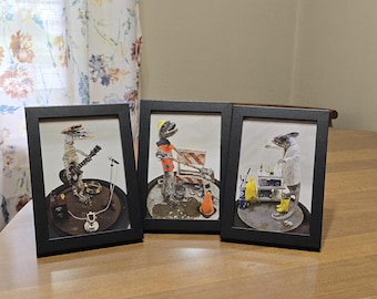 May include: Three framed art pieces with black frames. Each features a unique creature in a humorous setting: a lizard playing guitar, another operating construction equipment, and a third in a lab coat. The art is set on a brown surface.