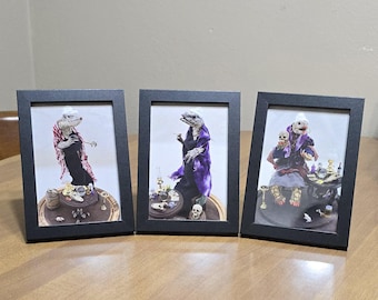 May include: Three framed photographs of lizard-like figures in various poses. The figures are dressed in purple and black robes and are holding various objects, including a goblet, a skull, and a crystal ball. The frames are black and have a simple design.