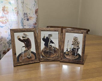 May include: Three framed art pieces featuring anthropomorphic figures using walkers. The frames are a dark wood grain. The figures are dressed in various outfits, with one wearing a brown sweater, another a black jacket, and the third a tan coat.