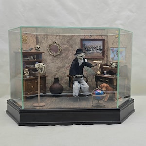 May include: A miniature diorama in a glass display case featuring a lizard-like figure in a black suit and hat sitting in a chair. The scene includes a wooden desk, a bookshelf with skulls, a globe, a small dog, and a map-patterned wall background.