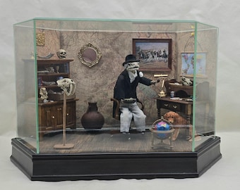 May include: A miniature diorama in a glass display case featuring a lizard-like figure in a black suit and hat sitting in a chair. The scene includes a wooden desk, a bookshelf with skulls, a globe, a small dog, and a map-patterned wall background.