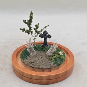 May include: A miniature diorama set in a round wooden base featuring two textured, pale, claw-like hands emerging from a sandy patch of ground. The scene includes a small dark cross, a sparse green tree, and patches of green moss on the surface.