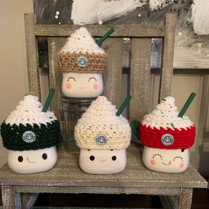 May include: Four white ceramic mugs with different coloured crocheted hats. The hats are decorated with a Starbucks logo and resemble a winter beverage. The mugs are sitting on a wooden shelf.