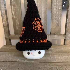 May include: A black crocheted witch hat with orange spiderweb accents sits on a white ceramic mug with a smiling face.