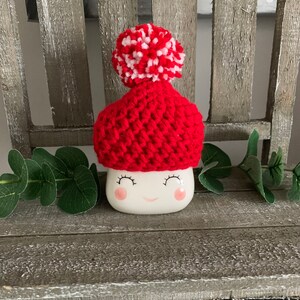 May include: White ceramic mug with a red and white knitted hat with a pom pom on top. The mug has a smiling face with pink cheeks and eyelashes.