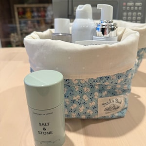May include: A light green cylindrical deodorant container labeled Salt & Stone Bergamot & Hinoki, positioned in front of a fabric storage basket. The basket features a blue and white floral pattern with a white interior and a Stitch & Bark label.