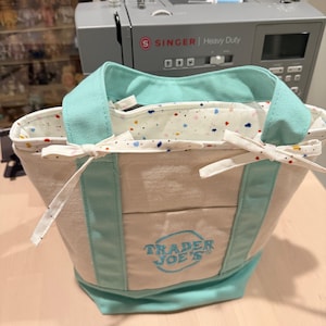 May include: A beige canvas tote bag with mint green handles and a matching base. The bag features an embroidered blue Trader Joe's logo on the front pocket and a white interior lining with a colorful confetti-like pattern, secured by two side ties.