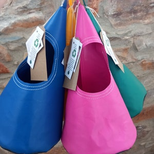 May include: Four hanging plant pouches in blue, pink, yellow, and green. The pouches are made of a water-resistant fabric and have a white stitched seam around the top. Each pouch has a small tag attached with the text "Sustainable & Reusable" and a green recycling symbol.