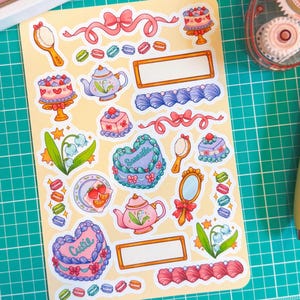 May include: A sheet of decorative stickers with a pastel theme. The stickers feature cakes, teapots, mirrors, bows, macarons, and floral elements. Some stickers have text that reads "Cutie" and "Smartle". The stickers are on a yellow background.