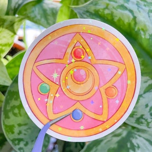 Peut inclure: Un autocollant rose et or avec une &eacute;toile &agrave; cinq branches et un croissant de lune au centre. L'&eacute;toile a un bijou rouge, vert et orange sur chaque pointe. L'autocollant a une finition holographique.