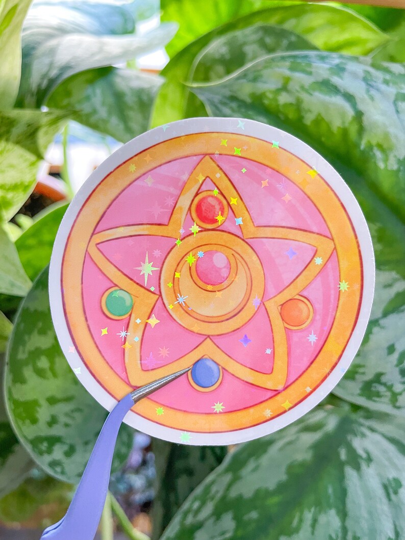 Peut inclure: Un autocollant rose et or avec une &eacute;toile &agrave; cinq branches et un croissant de lune au centre. L'&eacute;toile a un bijou rouge, vert et orange sur chaque pointe. L'autocollant a une finition holographique.