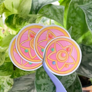 Peut inclure: Quatre autocollants roses et dor&eacute;s avec un motif d'&eacute;toile &agrave; cinq branches. L'&eacute;toile a un croissant de lune au centre et est entour&eacute;e d'un cercle de petites pierres pr&eacute;cieuses.