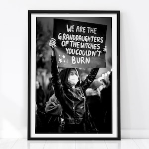 May include: A black and white framed print featuring a person in a leather jacket and face mask holding a sign that reads: WE ARE THE GRANDDAUGHTERS OF THE WITCHES YOU COULDN'T BURN. The sign includes small star illustrations and bold white text.