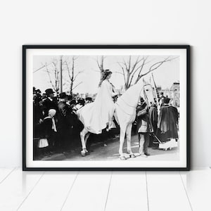 May include: Black and white framed print of a woman riding a white horse in a parade. The woman is wearing a long white dress and is surrounded by people in formal attire. The image is a vintage photograph.