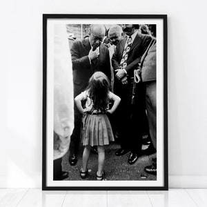May include: Black and white photograph of a young girl standing in front of a group of men. The girl is wearing a skirt and a blouse. The men are wearing suits and ties. The photo is framed in a black frame.
