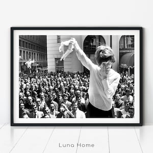 Black and white vintage photo of a young woman in 1969 smiling and holding up her bra defiantly in front of a large crowd of men, symbolizing feminist rebellion and the No Bra Movement.