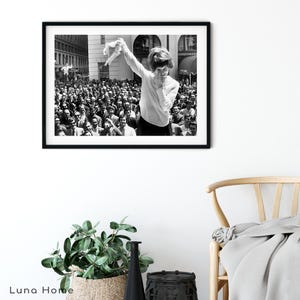 Puede incluir: Impresi&oacute;n enmarcada en blanco y negro que muestra a una mujer levantando una prenda frente a una gran multitud. La fotograf&iacute;a captura un momento de celebraci&oacute;n o triunfo, con una est&eacute;tica vintage. El fondo incluye edificios y una escena callejera.