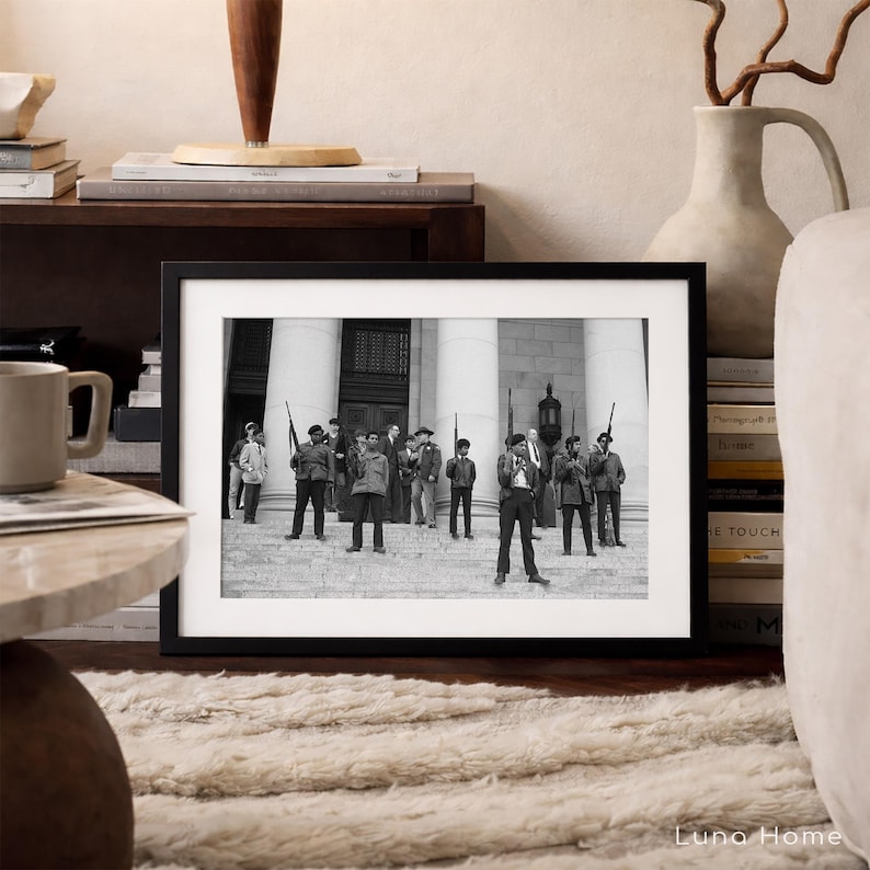 May include: A framed black and white photograph depicting a group of people, some holding rifles, standing on steps in front of a building with columns. The photograph is displayed on a wooden surface, with a mug and books nearby.