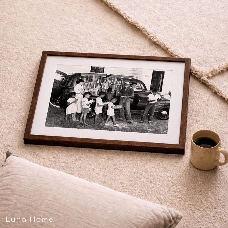 May include: A framed black and white photograph depicts a group of people gathered around a vehicle filled with books. The photograph is in a dark brown wooden frame, resting on a beige rug. A mug of coffee and a pillow are also in the image.