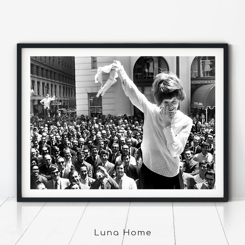 Black and white vintage photo of a young woman in 1969 smiling and holding up her bra defiantly in front of a large crowd of men, symbolizing feminist rebellion and the No Bra Movement.