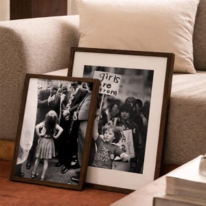 May include: Two framed black and white photographs with dark brown wooden frames. One shows a young girl standing in front of a crowd, and the other shows a child holding a sign that says "girls are strong". The frames are leaning against a sofa.