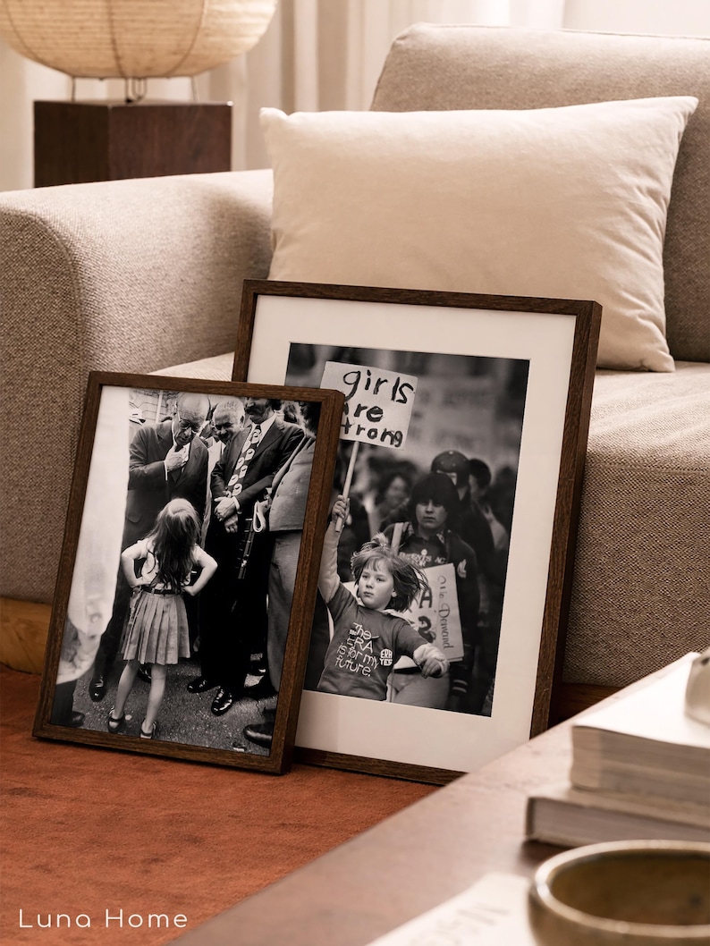 May include: Two framed black and white photographs with dark brown wooden frames. One shows a young girl standing in front of a crowd, and the other shows a child holding a sign that says "girls are strong". The frames are leaning against a sofa.