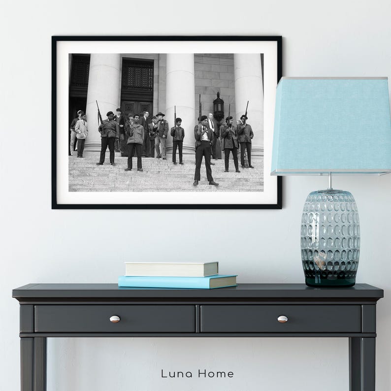 May include: Black and white framed photograph depicting a group of people standing on steps in front of a building with columns. The photograph is displayed above a black console table with a blue lamp.