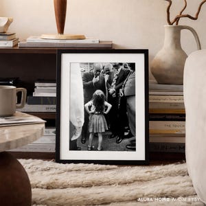 May include: A framed black and white photograph featuring a young girl standing with her hands on her hips, surrounded by a group of men in suits. The photograph is displayed on a shelf with books and decorative items. The text "ALURA HOME // ON ETSY" is visible.