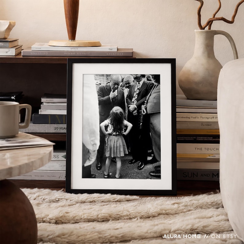 May include: A framed black and white photograph featuring a young girl standing with her hands on her hips, surrounded by a group of men in suits. The photograph is displayed on a shelf with books and decorative items. The text "ALURA HOME // ON ETSY" is visible.