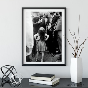 May include: Black and white framed photograph featuring a young girl standing with her hands on her hips, surrounded by a group of men in suits. The photograph is displayed on a black surface with decorative objects and books.