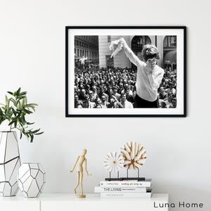 Puede incluir: Impresi&oacute;n enmarcada en blanco y negro de una mujer sonriendo y sosteniendo una prenda frente a una gran multitud. La imagen es una fotograf&iacute;a vintage, que captura un momento de celebraci&oacute;n. El marco negro contrasta con la pared blanca sobre la que se exhibe.