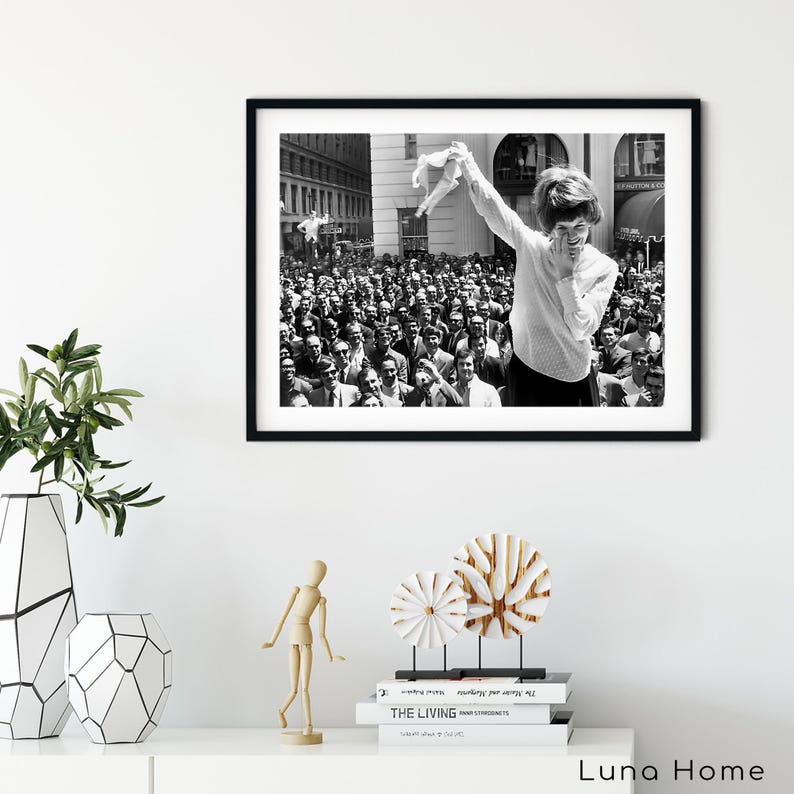 Puede incluir: Impresi&oacute;n enmarcada en blanco y negro de una mujer sonriendo y sosteniendo una prenda frente a una gran multitud. La imagen es una fotograf&iacute;a vintage, que captura un momento de celebraci&oacute;n. El marco negro contrasta con la pared blanca sobre la que se exhibe.