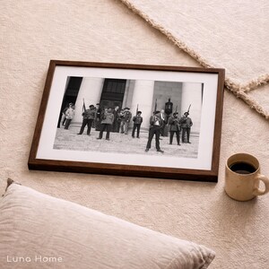 May include: A framed black and white photograph featuring a group of men in military attire, holding rifles, standing in front of a building with columns. The frame is dark brown, and a beige mug with coffee sits nearby.
