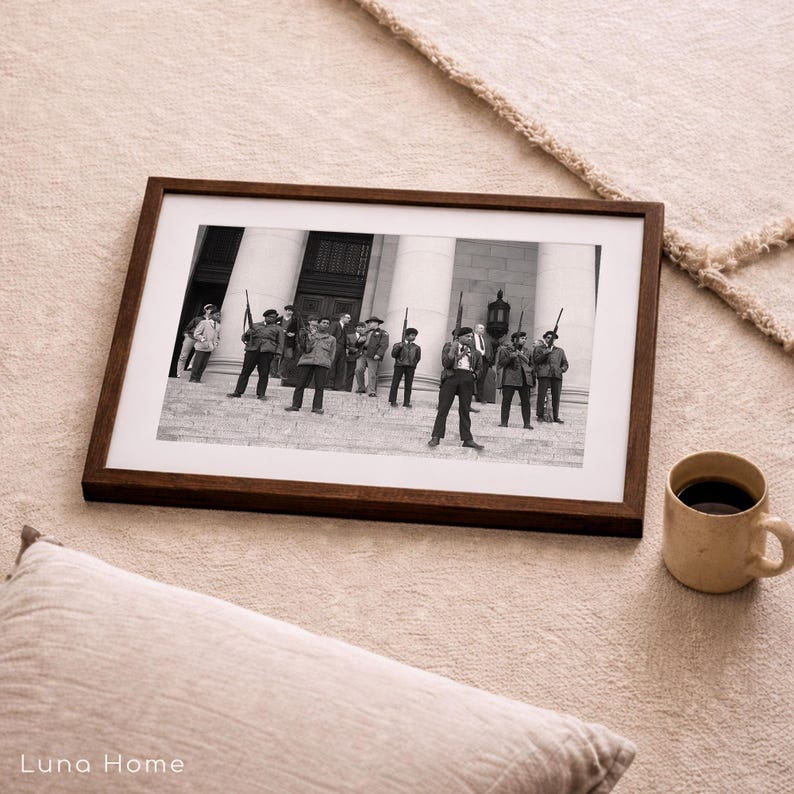 May include: A framed black and white photograph featuring a group of men in military attire, holding rifles, standing in front of a building with columns. The frame is dark brown, and a beige mug with coffee sits nearby.