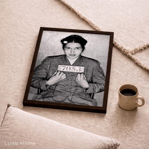 May include: A black and white framed portrait of Rosa Parks wearing a collared jacket and holding a sign with the number 7053. The rectangular frame is dark brown and surrounds the historical photograph, which shows a front-facing view.