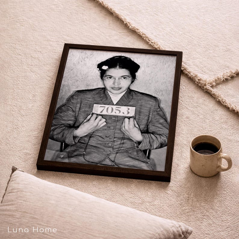 May include: A black and white framed portrait of Rosa Parks wearing a collared jacket and holding a sign with the number 7053. The rectangular frame is dark brown and surrounds the historical photograph, which shows a front-facing view.