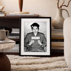 May include: A black and white framed portrait of Rosa Parks in a mugshot style, wearing a collared jacket and holding a sign with the number 7053. The print is displayed in a simple black frame, capturing a historical moment in a clear, high-contrast monochromatic tone.