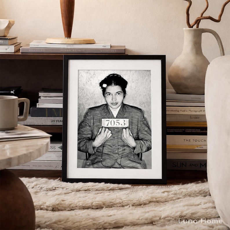 May include: A black and white framed portrait of Rosa Parks in a mugshot style, wearing a collared jacket and holding a sign with the number 7053. The print is displayed in a simple black frame, capturing a historical moment in a clear, high-contrast monochromatic tone.