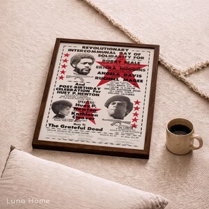 Pu&ograve; includere: Un poster incorniciato con una cornice in legno marrone, con testo e immagini relative a un evento politico. Il poster include il testo "Revolutionary Intercommunal Day of Solidarity" e i nomi Bobby Seale, Ericka Huggins, Angela Davis e Ruchell Magee.