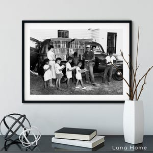 May include: Black and white framed print of a vintage scene. A group of children and adults are gathered around a vehicle filled with books, likely a mobile library. The image captures a moment of community and learning.