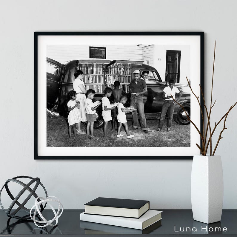 May include: Black and white framed print of a vintage scene. A group of children and adults are gathered around a vehicle filled with books, likely a mobile library. The image captures a moment of community and learning.
