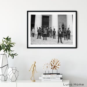 May include: Black and white framed photograph of a group of people standing on steps in front of a building. The people are holding rifles. The photograph is displayed on a white wall above a white table with decorative items.