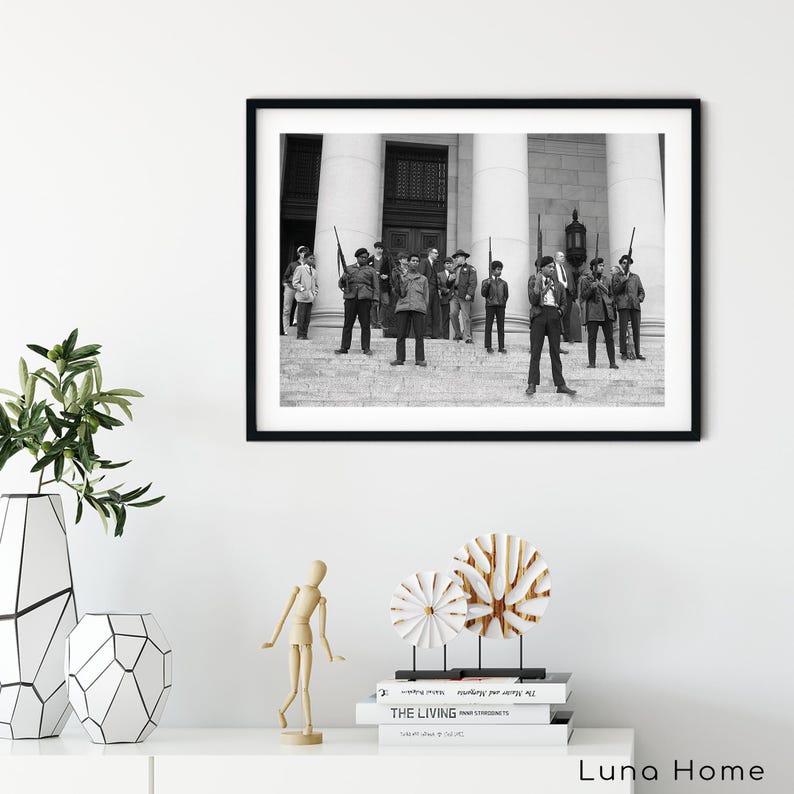 May include: Black and white framed photograph of a group of people standing on steps in front of a building. The people are holding rifles. The photograph is displayed on a white wall above a white table with decorative items.