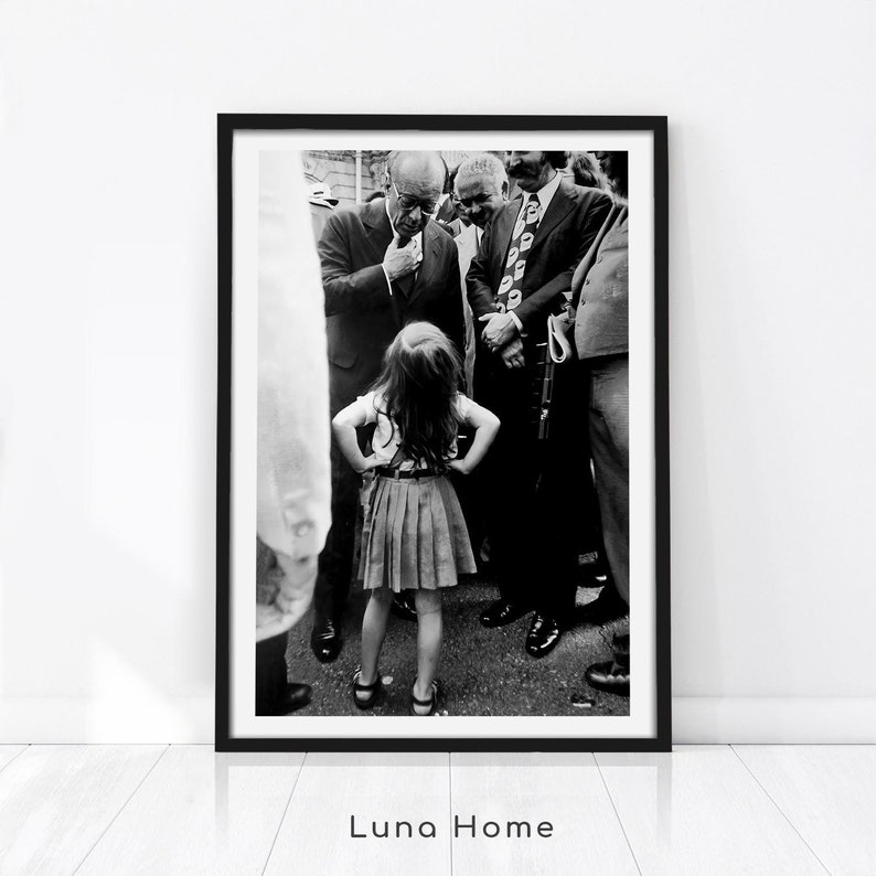 May include: Black and white photograph of a young girl standing in front of a group of men. The girl is wearing a skirt and a blouse. The men are wearing suits and ties. The photo is framed in a black frame.