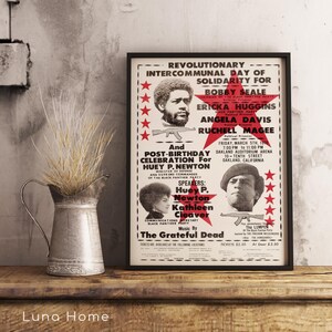 Pu&ograve; includere: Un poster incorniciato con bordo nero, con ritratti e testo. Il poster commemora una "Giornata Intercomunitaria Rivoluzionaria di Solidariet&agrave;" con nomi come Bobby Seale e Angela Davis. Una brocca di metallo con piante secche si trova a sinistra.