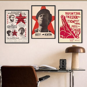 Pu&ograve; includere: Tre poster incorniciati con cornici nere sono esposti su un muro. I poster presentano testo e immagini di personaggi politici. Un poster ha il testo "Huey P. Newton". Una scrivania con una lampada e una fotocamera &egrave; di fronte ai poster.